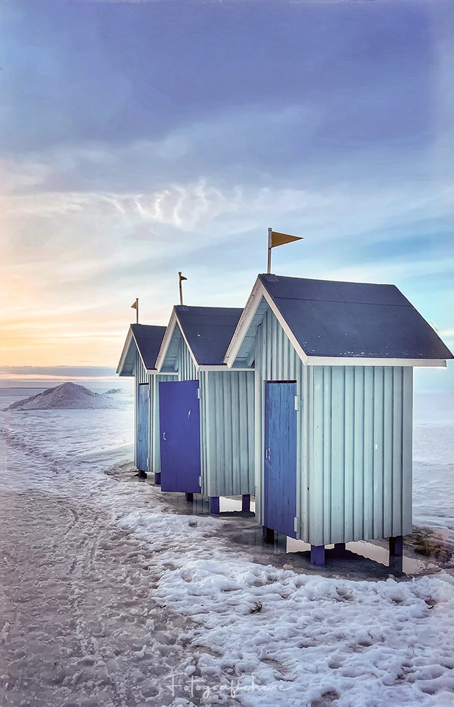 Drei blau-weiße Strandhäuschen mit dunklen Dächern am verschneiten Nallikari Strand in Oulu, Finnland, unter einem pastellfarbenen Abendhimmel.