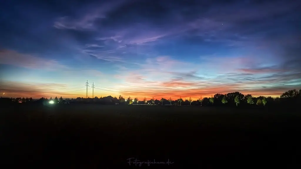 Farbenprächtiger Sonnenuntergang mit orange-rotem Horizont, türkisfarbenem und violettem Himmel, zarten Wolken, schmaler Mondsichel und dunklen Silhouetten von Bäumen und Strommasten