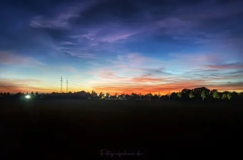 Farbenprächtiger Sonnenuntergang mit orange-rotem Horizont, türkisfarbenem und violettem Himmel, zarten Wolken, schmaler Mondsichel und dunklen Silhouetten von Bäumen und Strommasten