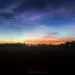 Farbenprächtiger Sonnenuntergang mit orange-rotem Horizont, türkisfarbenem und violettem Himmel, zarten Wolken, schmaler Mondsichel und dunklen Silhouetten von Bäumen und Strommasten