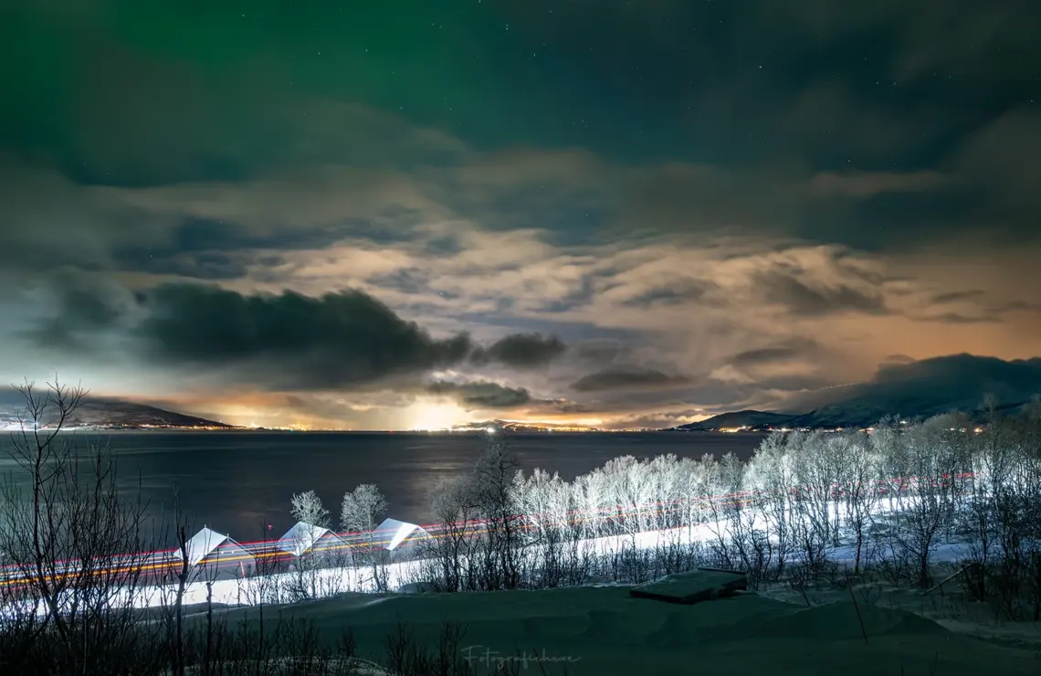 Nächtlicher Blick über den Fjord nach Tromsø mit grünen Polarlichtern, Wolken, den Lichtern eines landenden Flugzeugs und Autolichtspuren im Schnee.