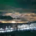 Nächtlicher Blick über den Fjord nach Tromsø mit grünen Polarlichtern, Wolken, den Lichtern eines landenden Flugzeugs und Autolichtspuren im Schnee.